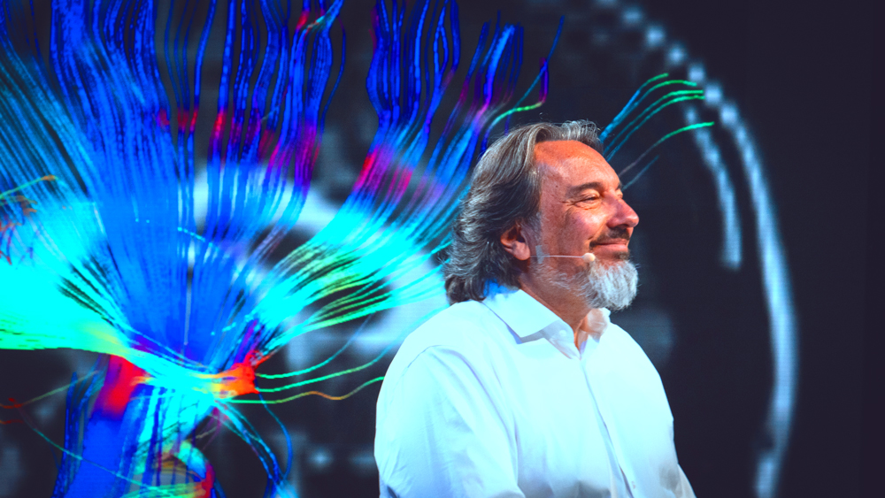 Patrizio Paoletti sorride davanti a un'immagine del cervello con le fibre nervose in blu e ciano